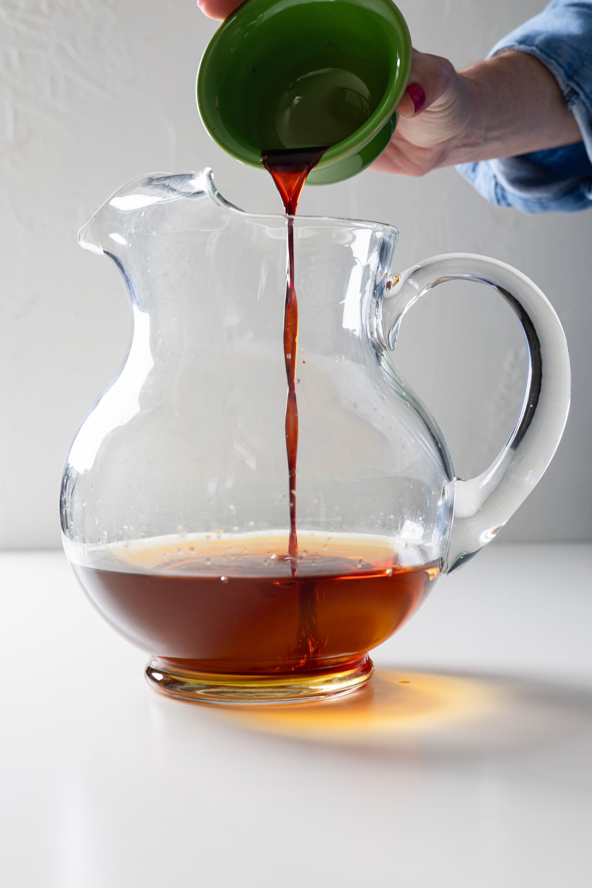 A hand pours a dark liquid from a small green bowl into a large clear glass pitcher containing more of the same large batch old fashioned, set on a white surface.