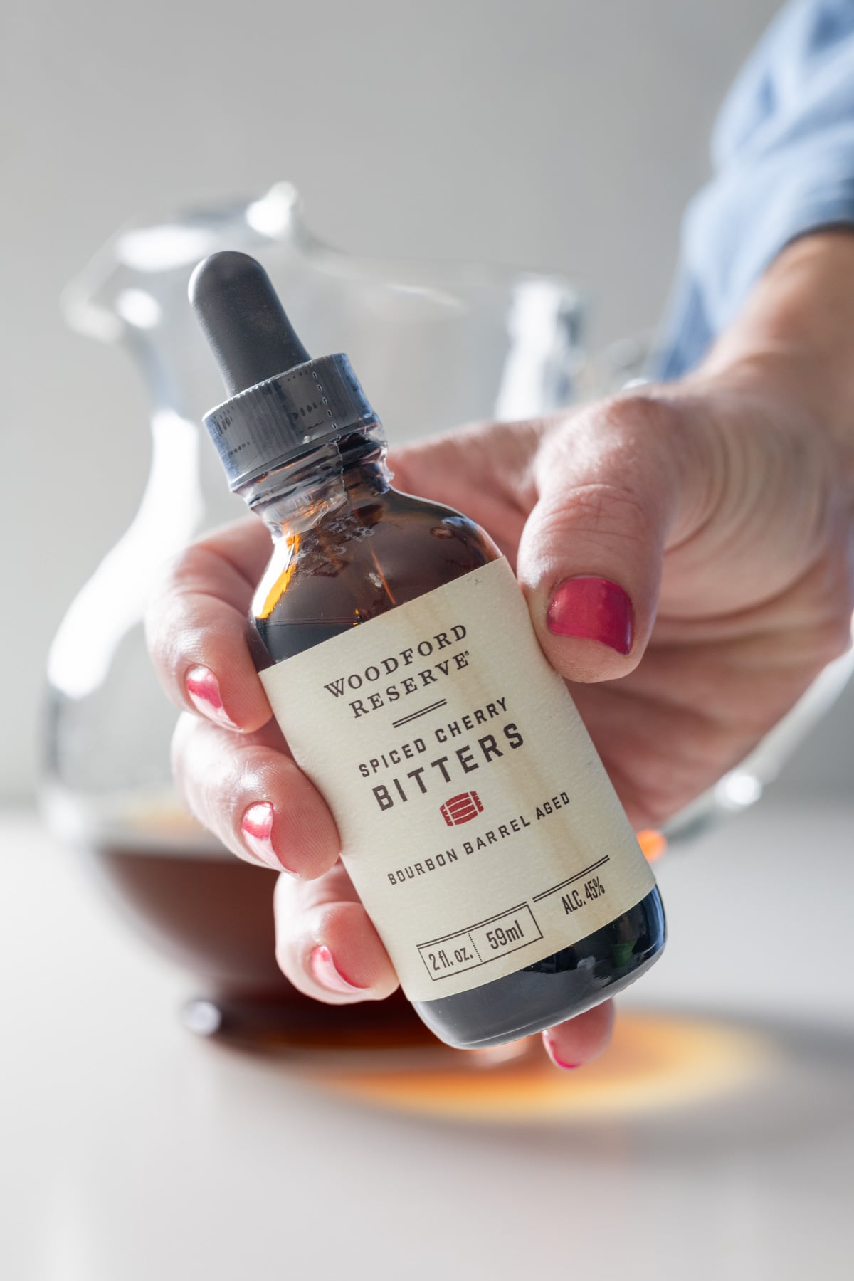 A hand with pink nail polish holds a bottle of Woodford Reserve Spiced Cherry Bitters with a dropper. In the background, a glass pitcher filled with a dark liquid hints at a large batch old fashioned ready to serve.