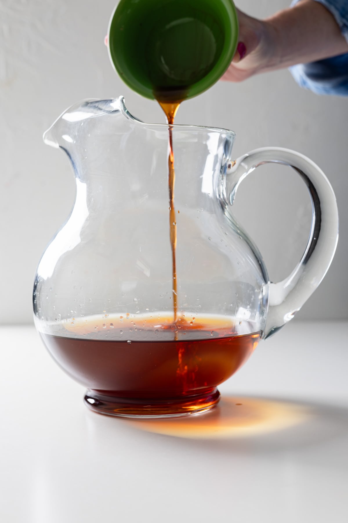 A hand pours a stream of dark liquid from a green cup into a clear glass pitcher, preparing a large batch old fashioned, all set against a plain white background.