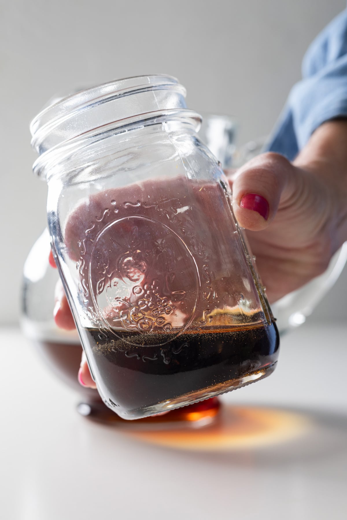 A hand with red nail polish holds a glass mason jar containing a small amount of dark liquid, possibly coffee or a large batch old fashioned, with more coffee visible in a glass container in the background.