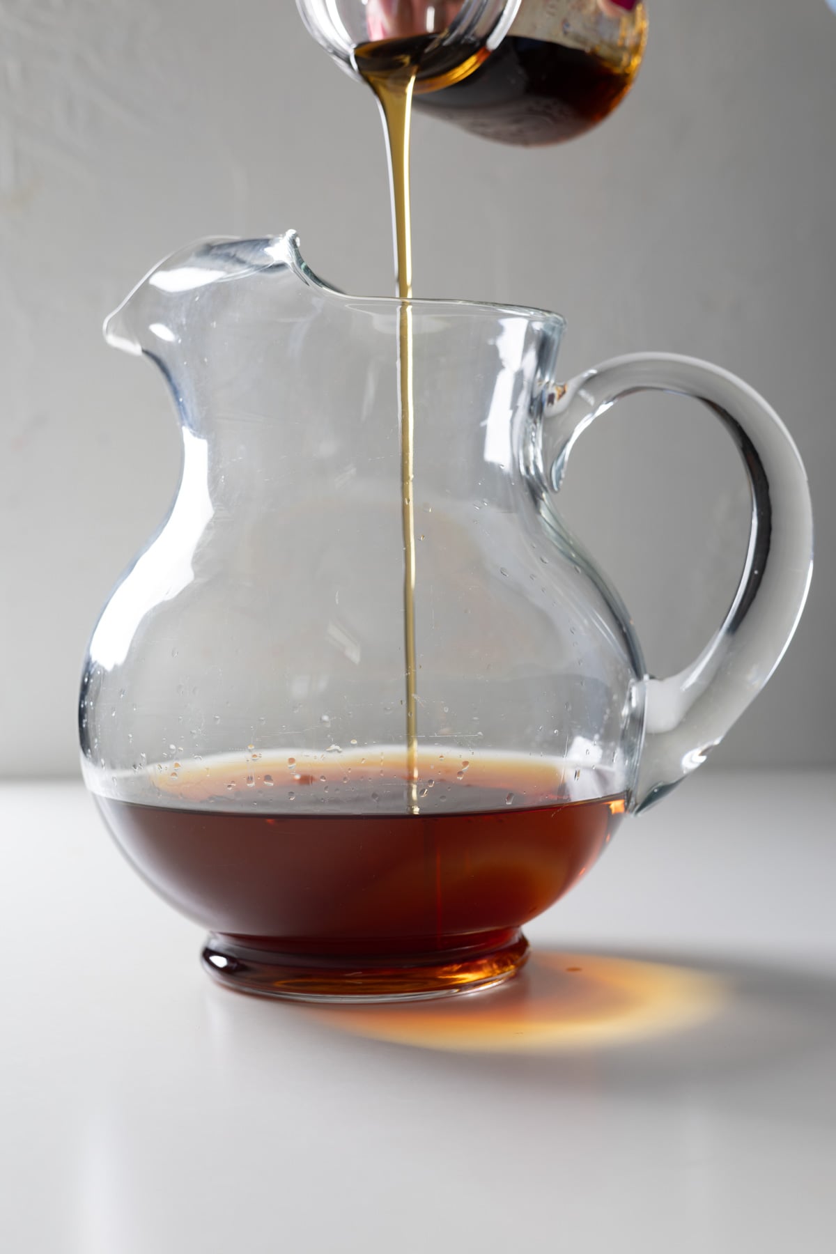 A stream of syrup is being poured from a container into a clear glass pitcher, partially filled with syrup, perfect for crafting a large batch old fashioned on the pristine white surface.