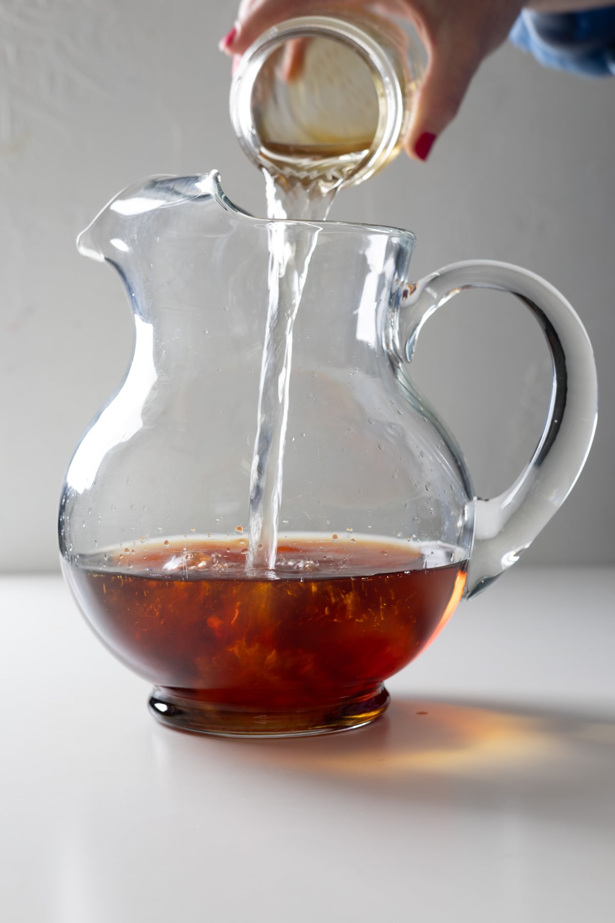 A hand pours a clear liquid from a small glass jar into a glass pitcher containing a dark amber large batch old fashioned, set on a white surface with a light background.