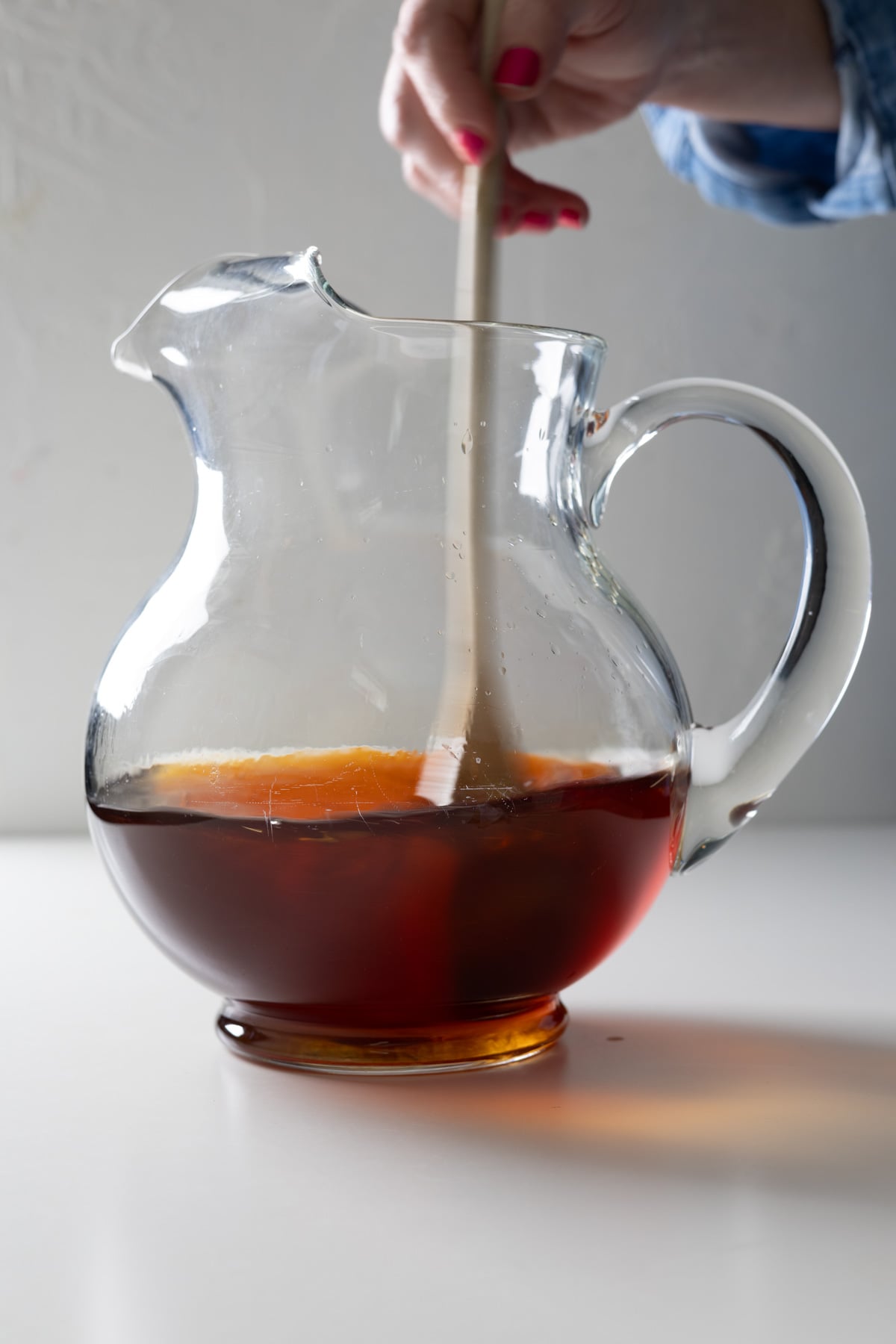 A hand with red nail polish stirs a large batch old fashioned with a wooden spoon in a clear glass pitcher on a white surface.