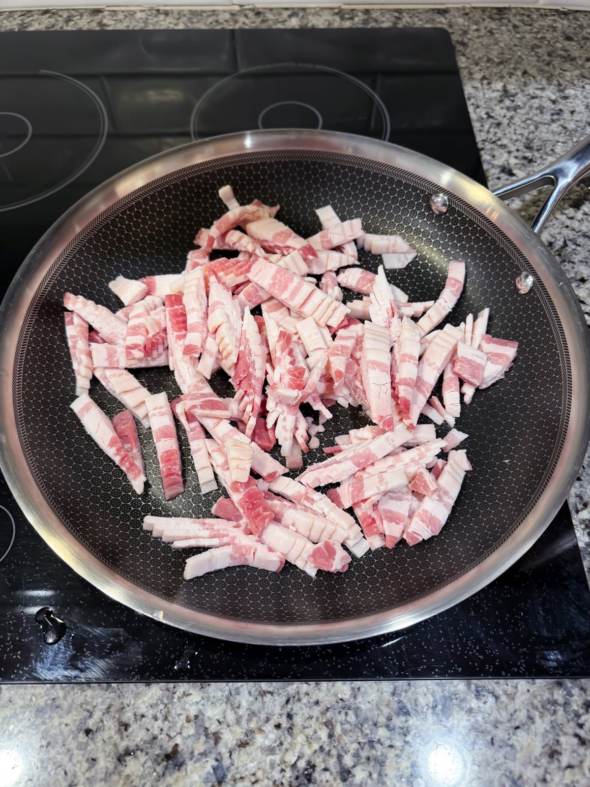 A frying pan on a stovetop contains uncooked, sliced strips of bacon arranged unevenly across the cooking surface. The pan rests on a granite countertop.