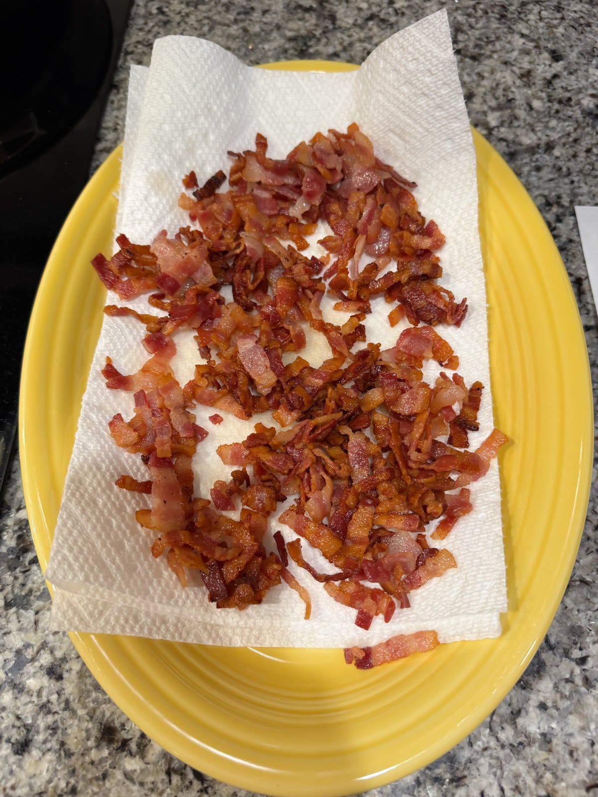 Chopped cooked bacon pieces are spread on a paper towel, which sits on a yellow oval plate. The plate is placed on a speckled granite countertop.