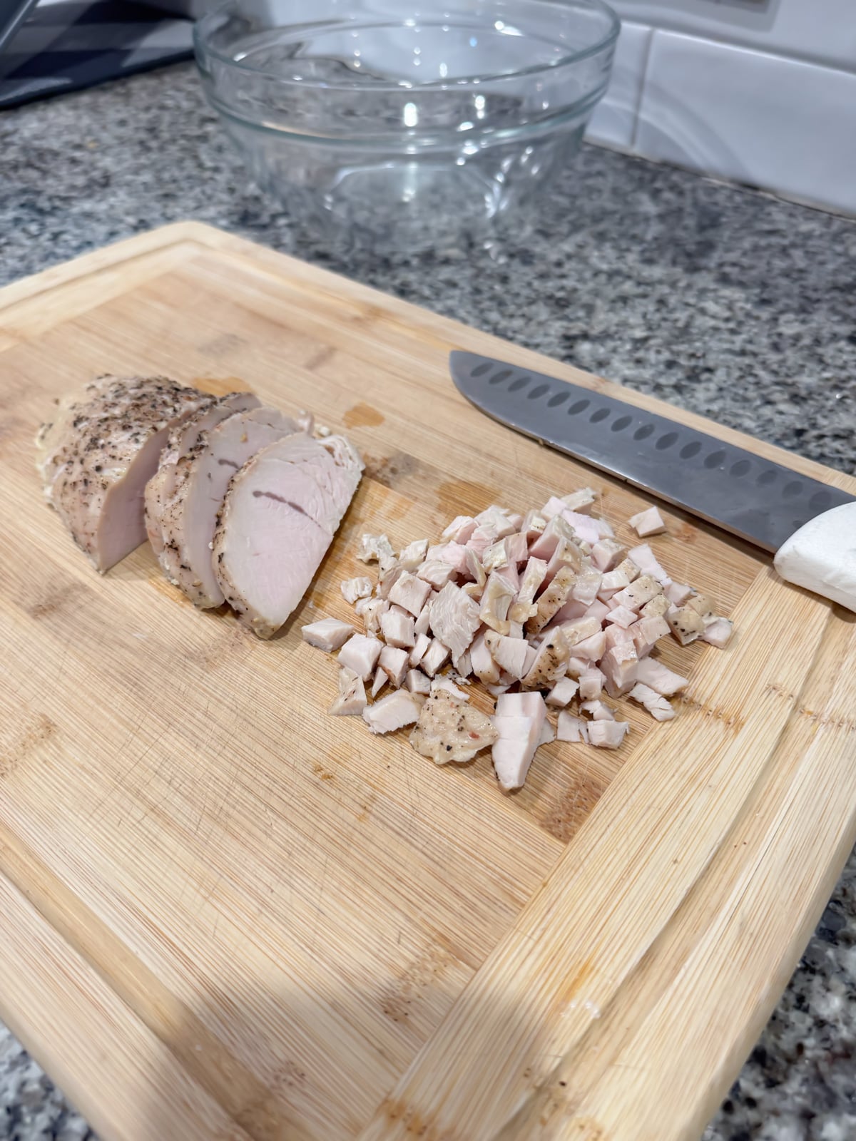Sliced and diced cooked pork on a wooden cutting board with a chefs knife, set on a kitchen countertop next to a glass mixing bowl.
