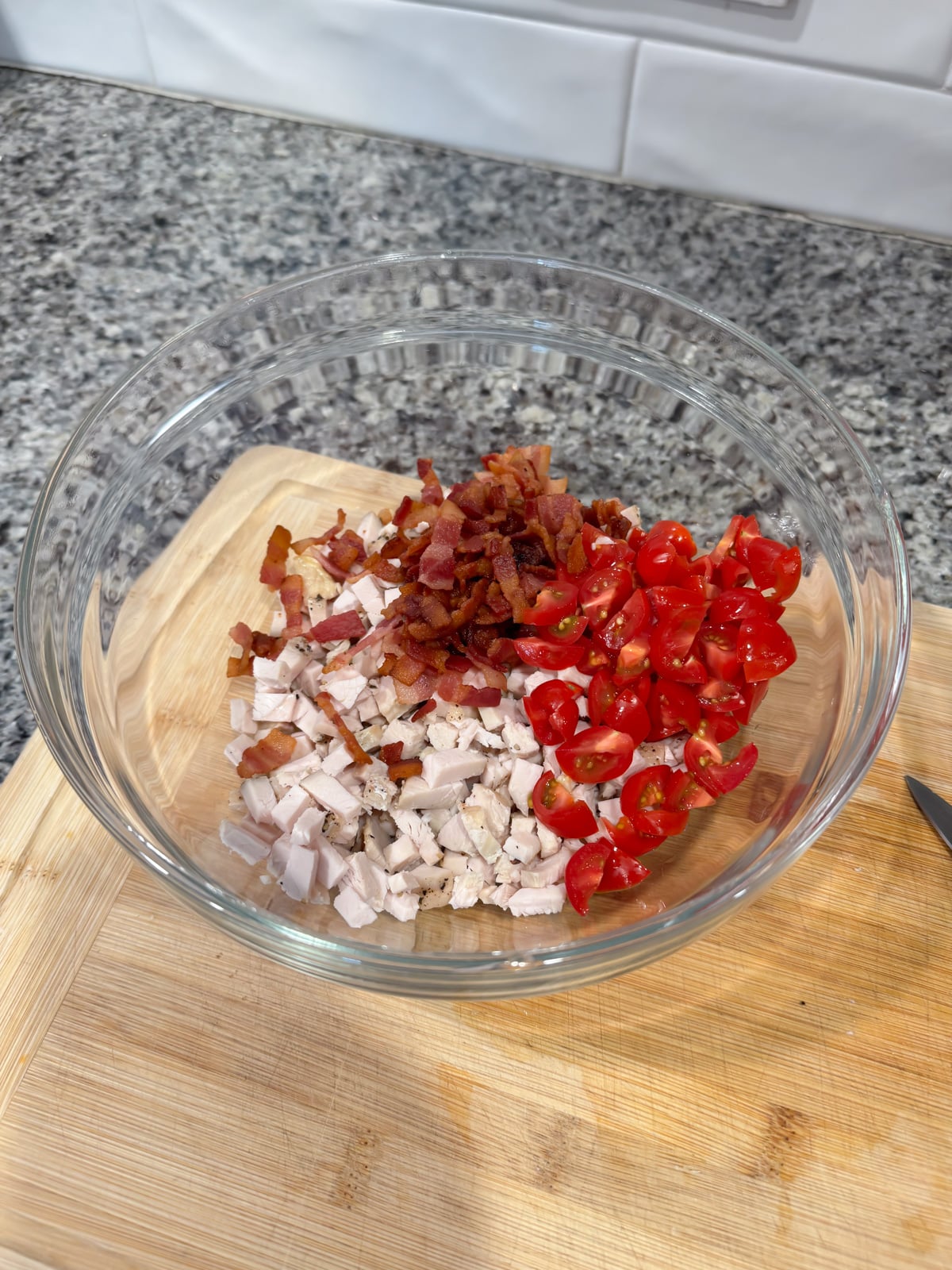 A glass bowl on a wooden cutting board contains chopped cooked bacon, diced turkey, and halved cherry tomatoes, arranged in separate sections. The background shows a granite countertop and white tile backsplash.
