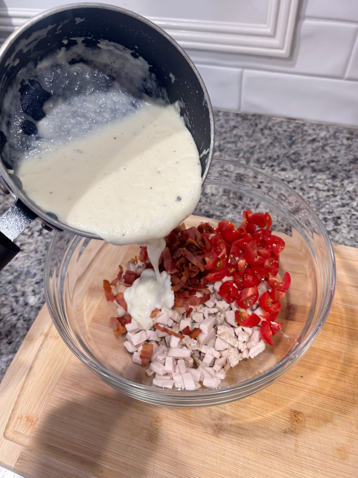 A saucepan pours creamy white sauce into a glass bowl containing chopped cooked chicken, bacon bits, and diced cherry tomatoes on a wooden cutting board.