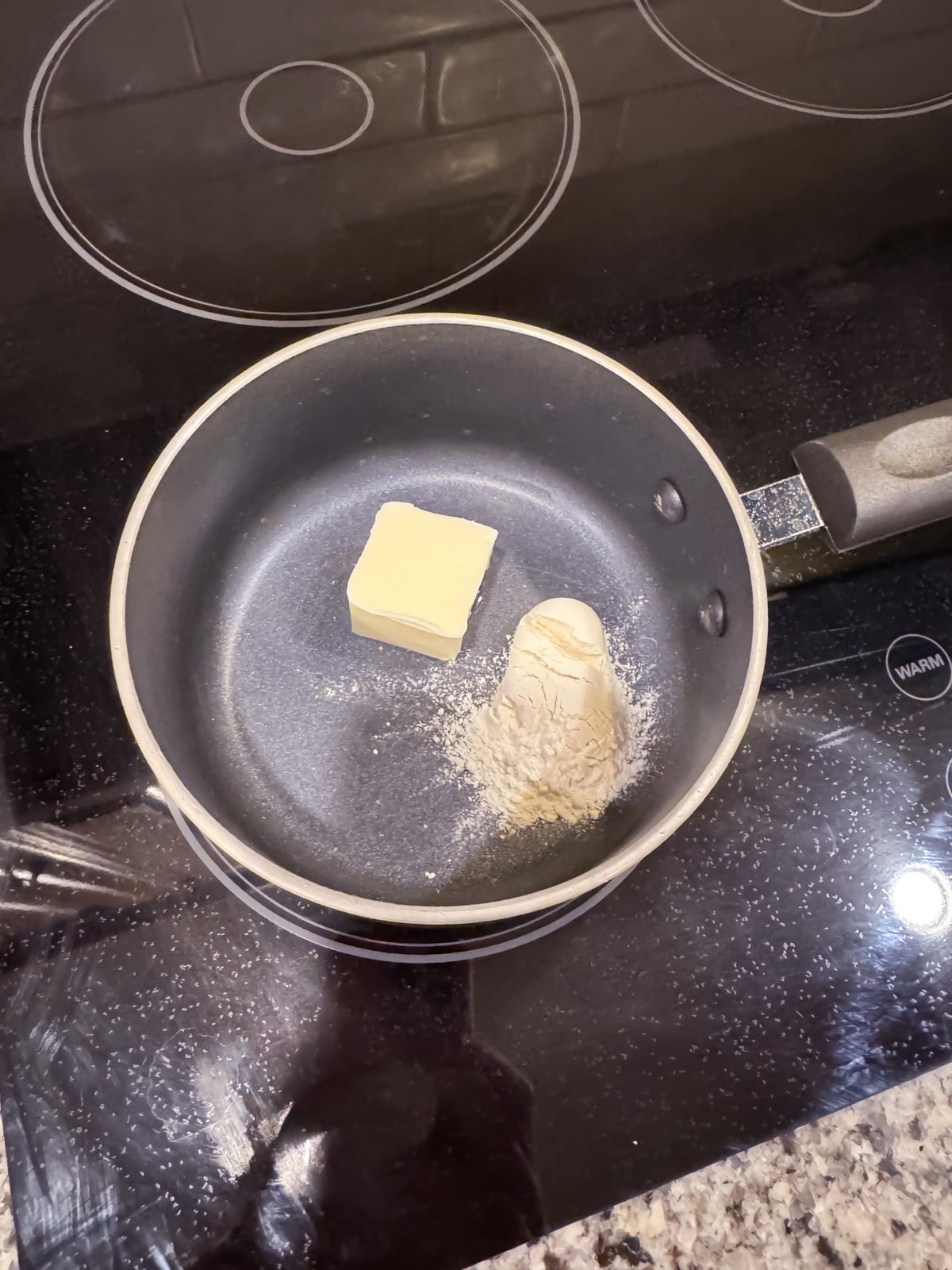 A small saucepan on a stovetop contains a pat of butter and a pile of flour, with more flour scattered on the stove and nearby countertop.