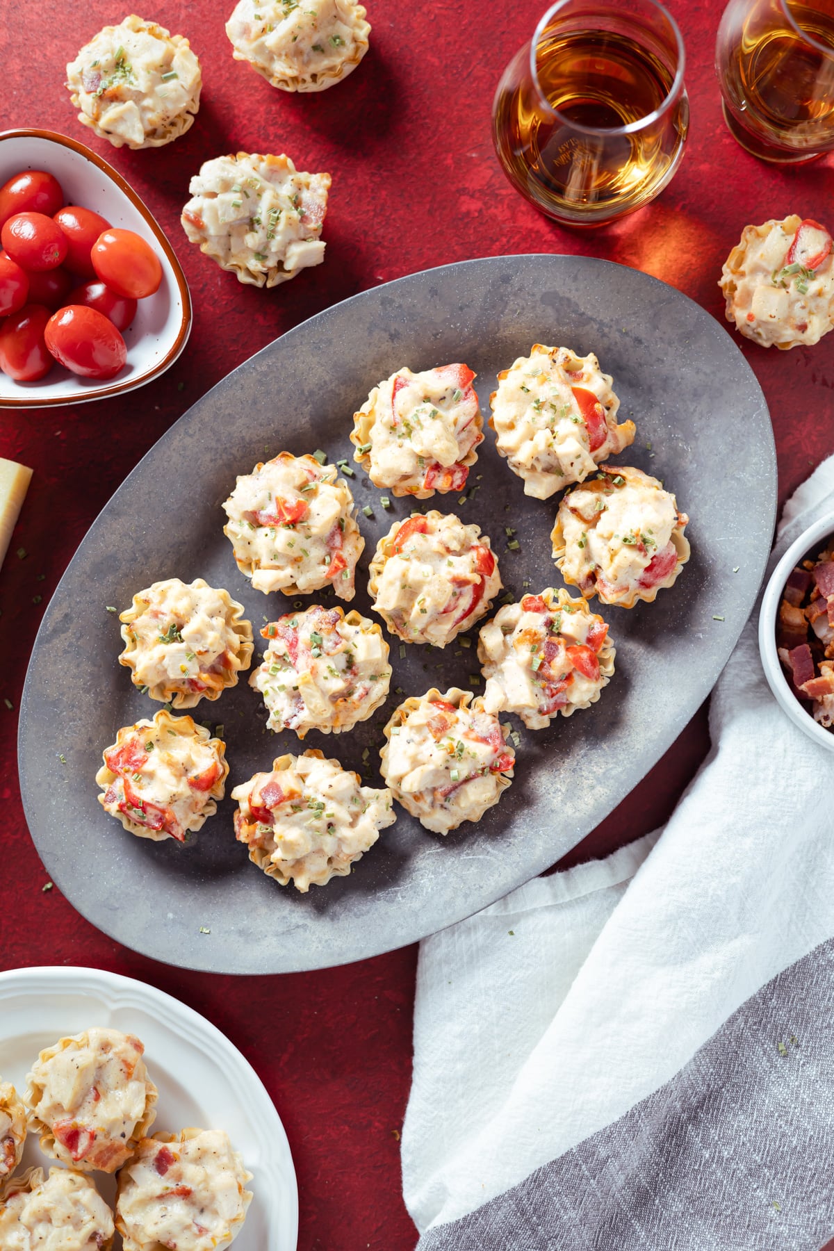 mini hot brown bites on a silver platter on a red background. glasses of bourbon, tomatoes, and plates of hot brown bites around it.