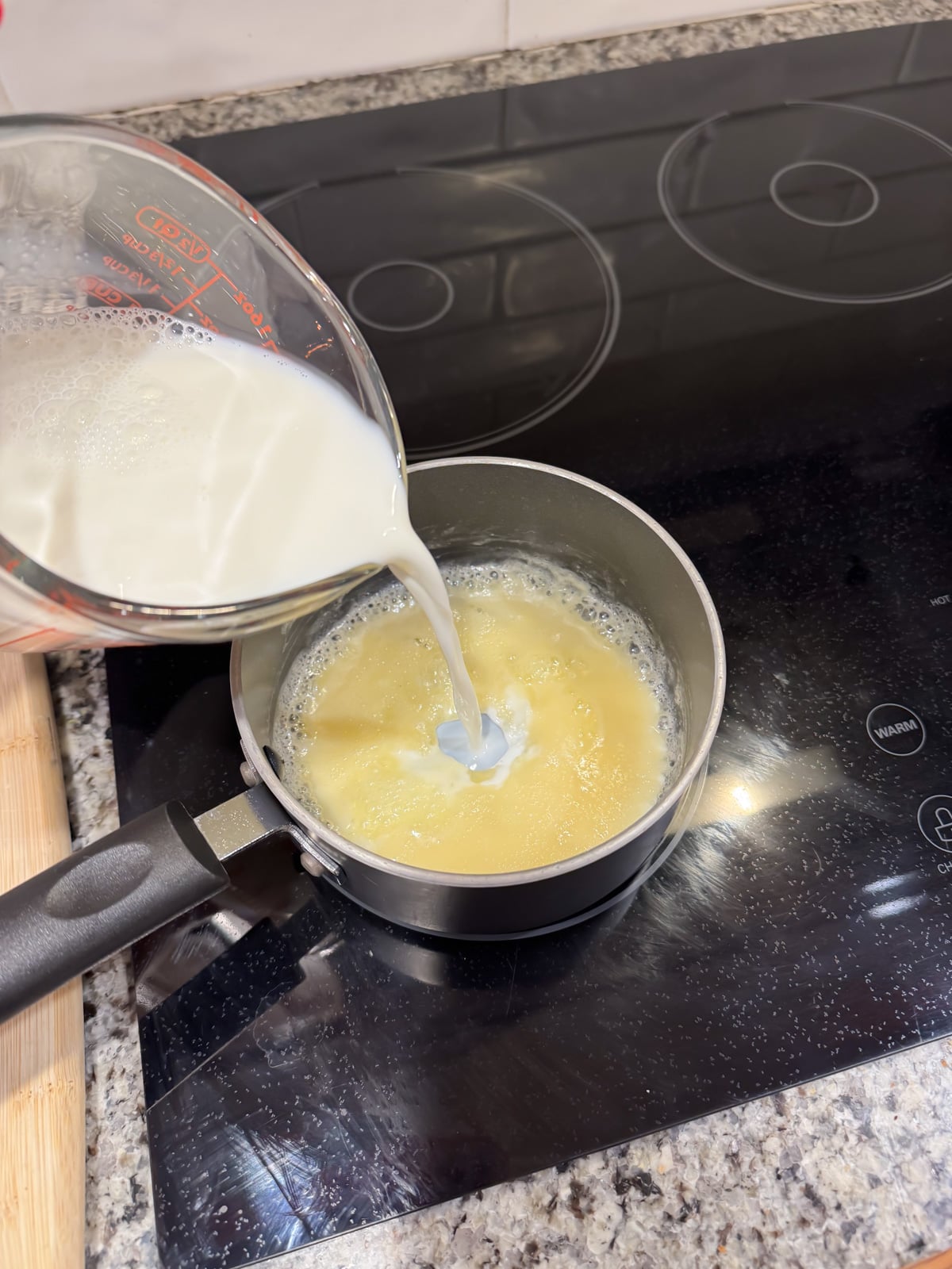 A hand pours milk from a measuring cup into a saucepan containing a yellow mixture, on a black electric stovetop with a granite countertop.
