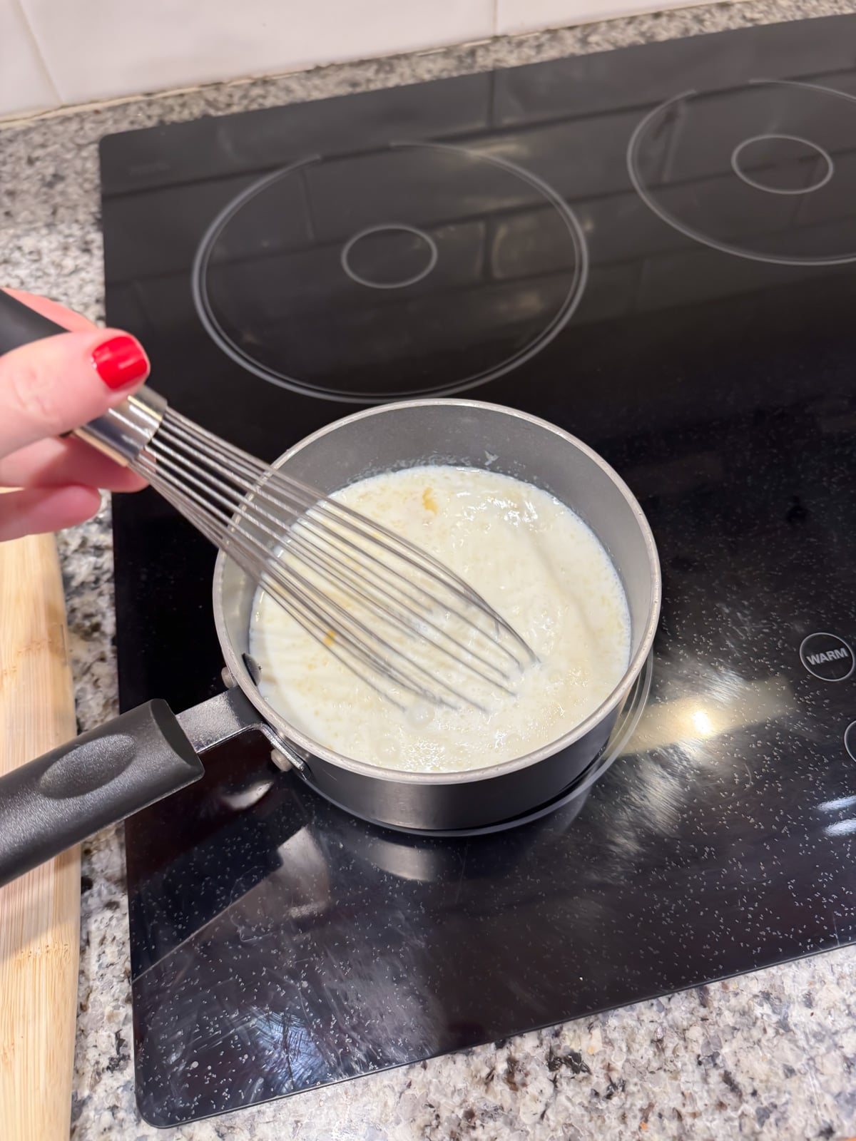 A hand with red nail polish is whisking a creamy mixture in a small saucepan on a black electric stovetop. The saucepan is on a granite countertop next to a wooden cutting board.