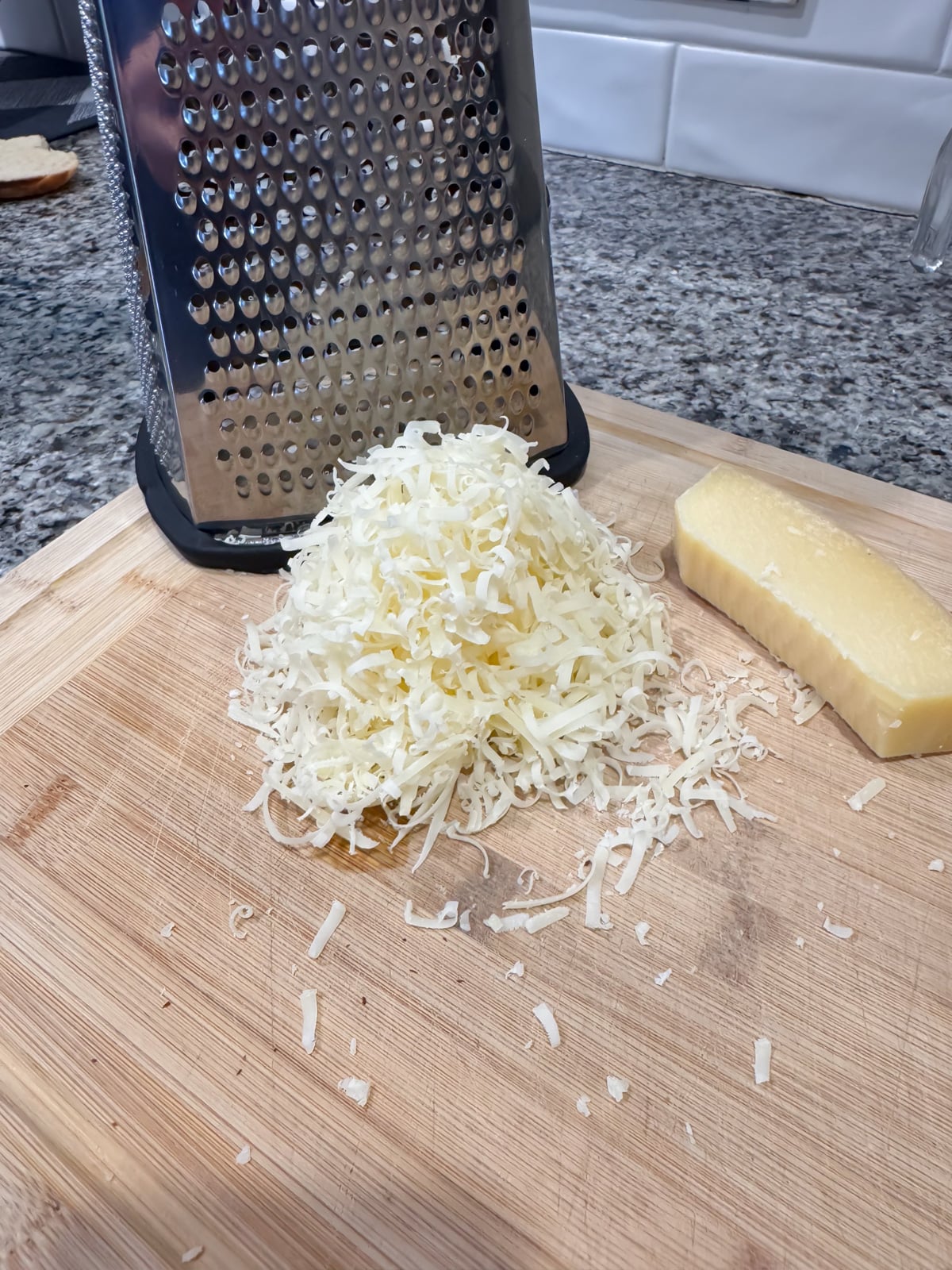 A pile of freshly grated cheese sits on a wooden cutting board beside a metal box grater and a block of cheese, on a granite countertop with white subway tile backsplash.