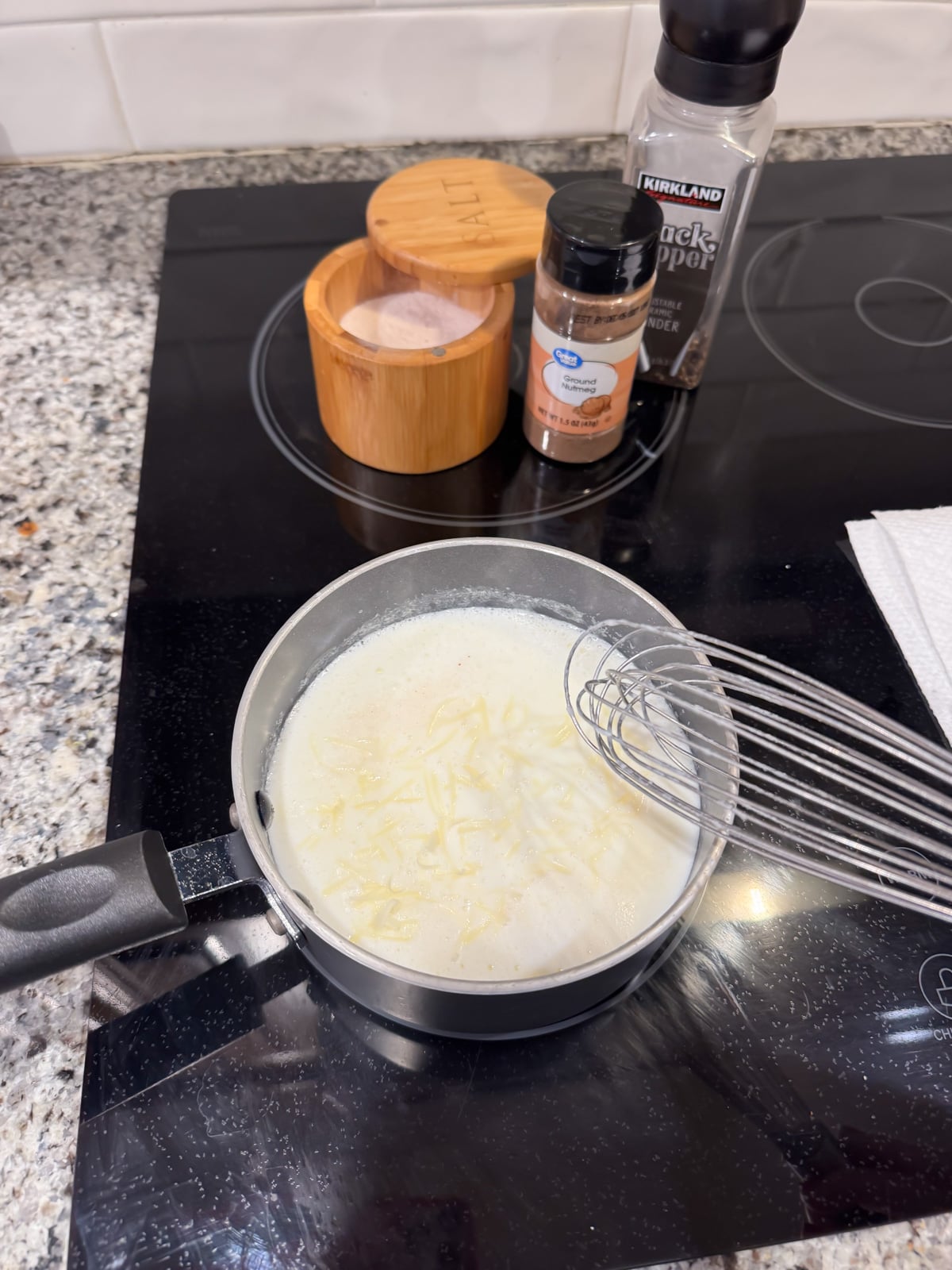 A small saucepan with a creamy white sauce and shredded cheese being whisked on a stovetop. Nearby are containers of salt, black pepper, and ground nutmeg on a granite countertop.