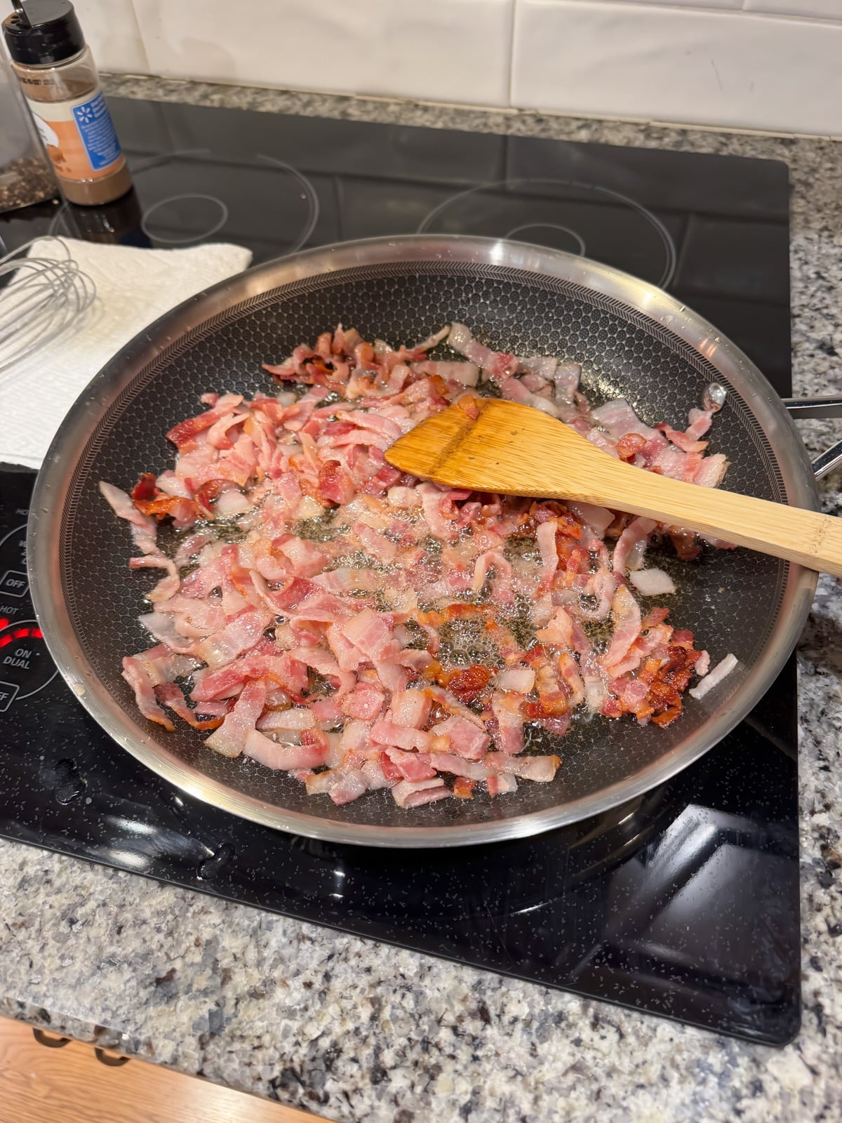 A frying pan on a stovetop with chopped bacon sizzling and being stirred with a wooden spatula; a whisk, seasoning bottle, and paper towel are nearby on the granite countertop.