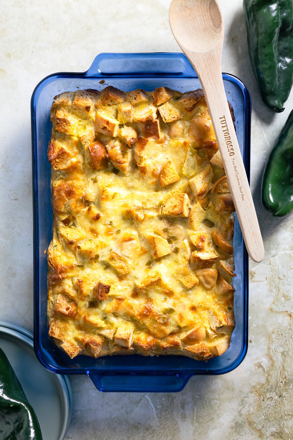 A baked casserole with golden, cubed bread pieces in a blue dish, topped with melted cheese. A wooden spoon rests on the dish. Green poblano peppers and blue plates are nearby on a light surface.
