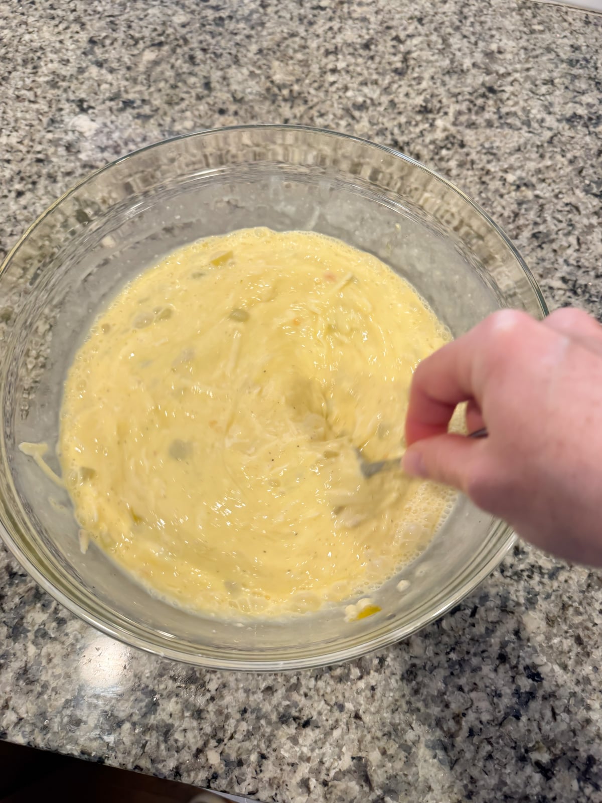 A hand stirring eggs, milk, and spices together in a large mixing bowl on a counter top.