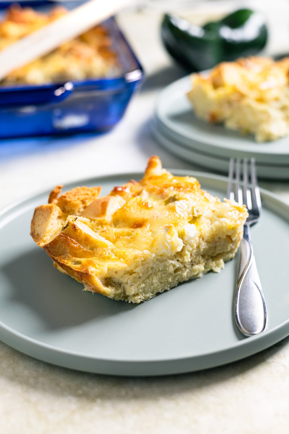 A slice of savory breakfast casserole with a golden top is served on a light gray plate with a fork. Another plate and a blue baking dish with more pudding are visible in the background.