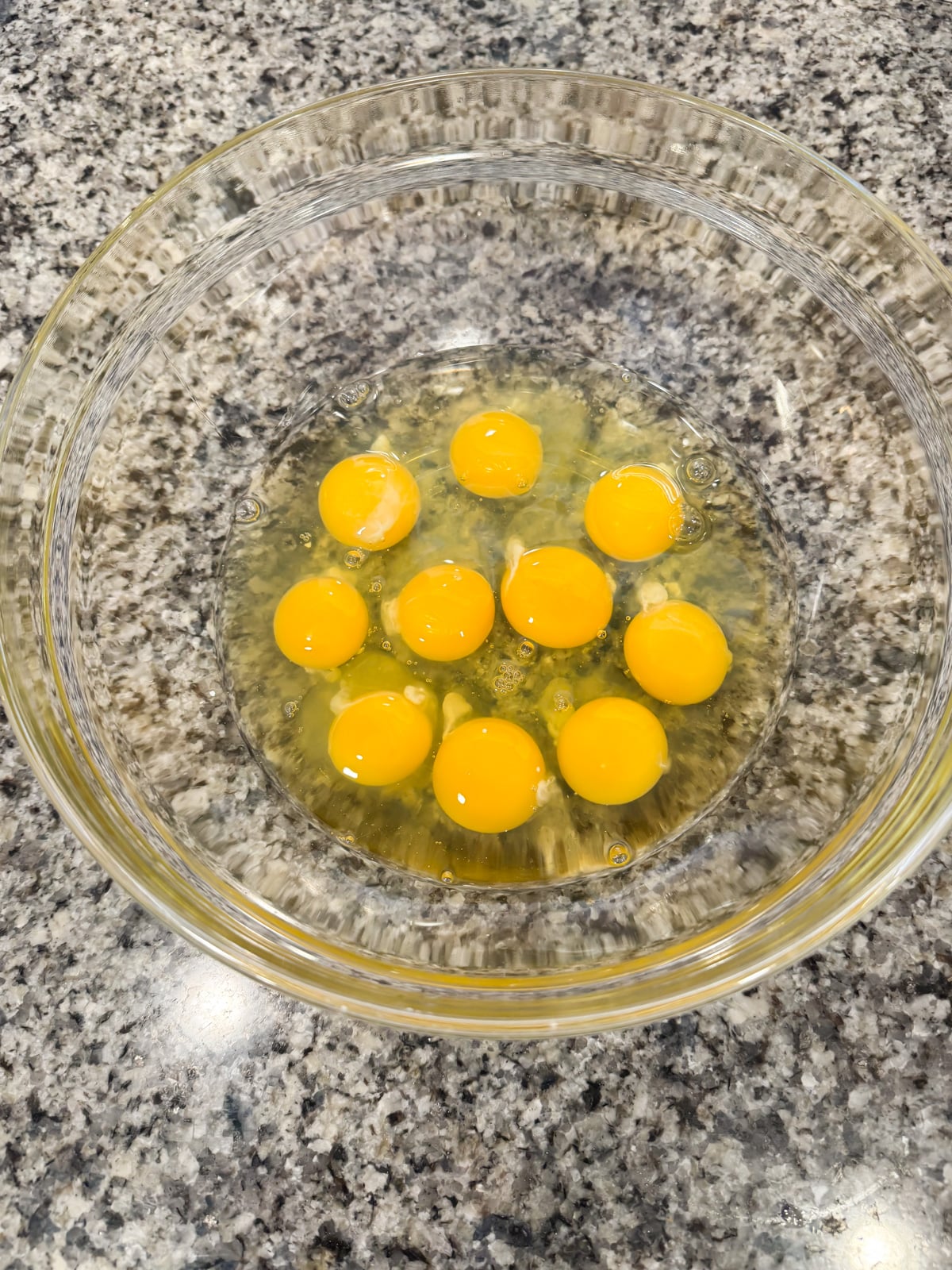 A clear glass bowl containing ten raw eggs with intact yolks, sitting on a speckled granite countertop.