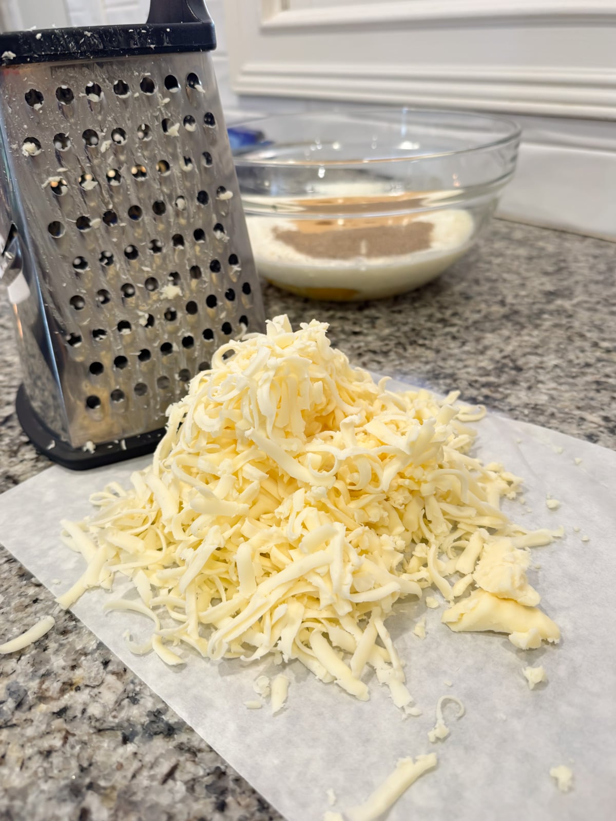 A pile of freshly grated cheese sits on parchment paper next to a metal box grater on a kitchen counter, with a glass mixing bowl containing dry ingredients in the background.