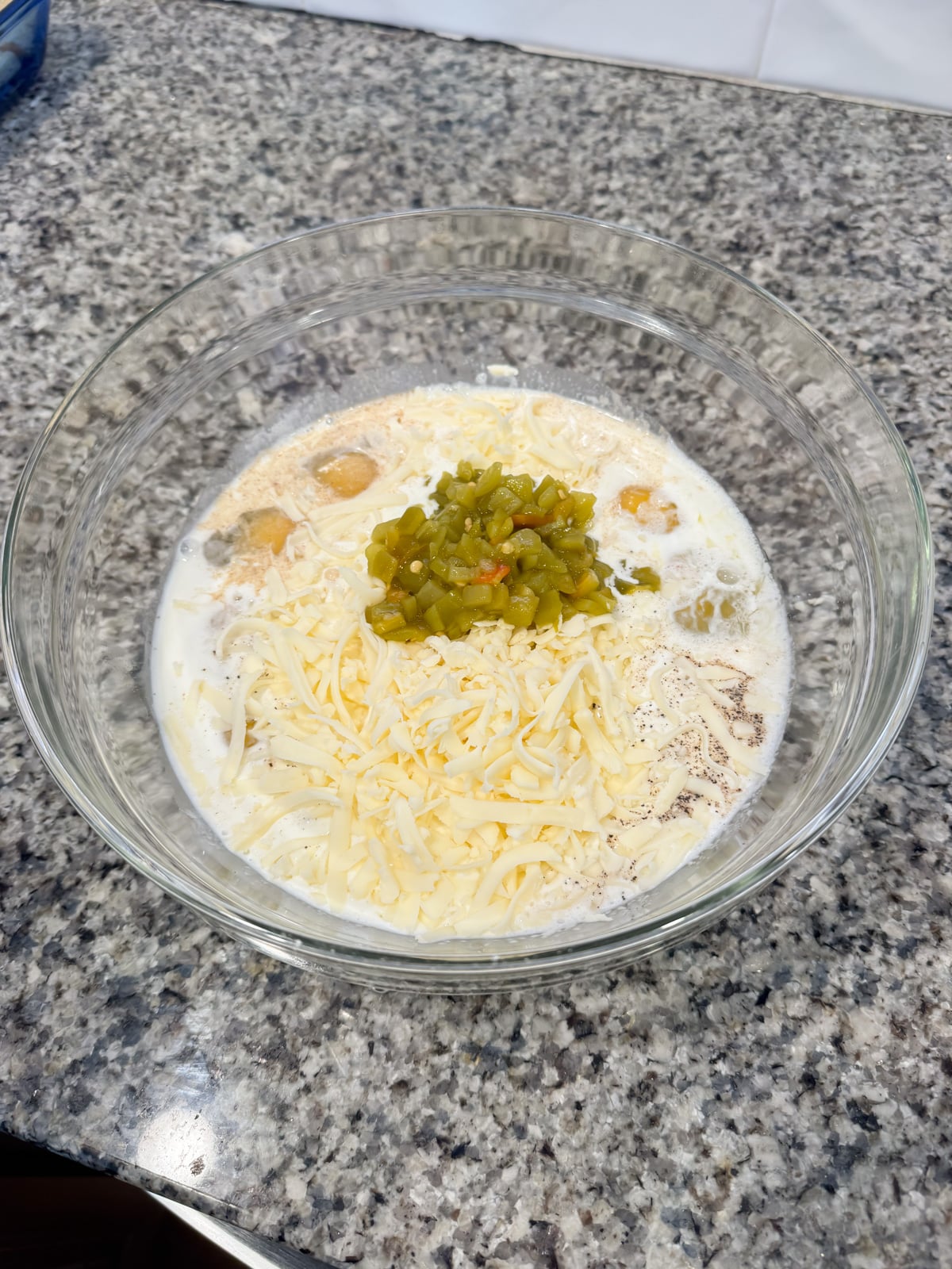 A glass bowl on a granite countertop containing shredded cheese, chopped green chiles, tater tots, milk, and seasonings, ready to be mixed together.