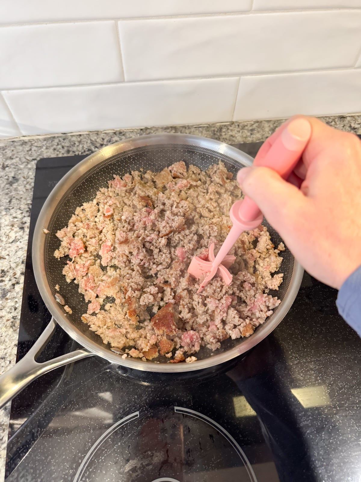 Breaking up sausage with a utensil as it's cooking in a large skillet.