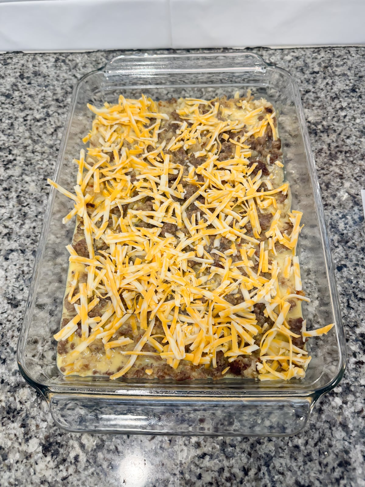 Shredded cheese on top of hash brown casserole in a baking pan before going into the oven to bake.