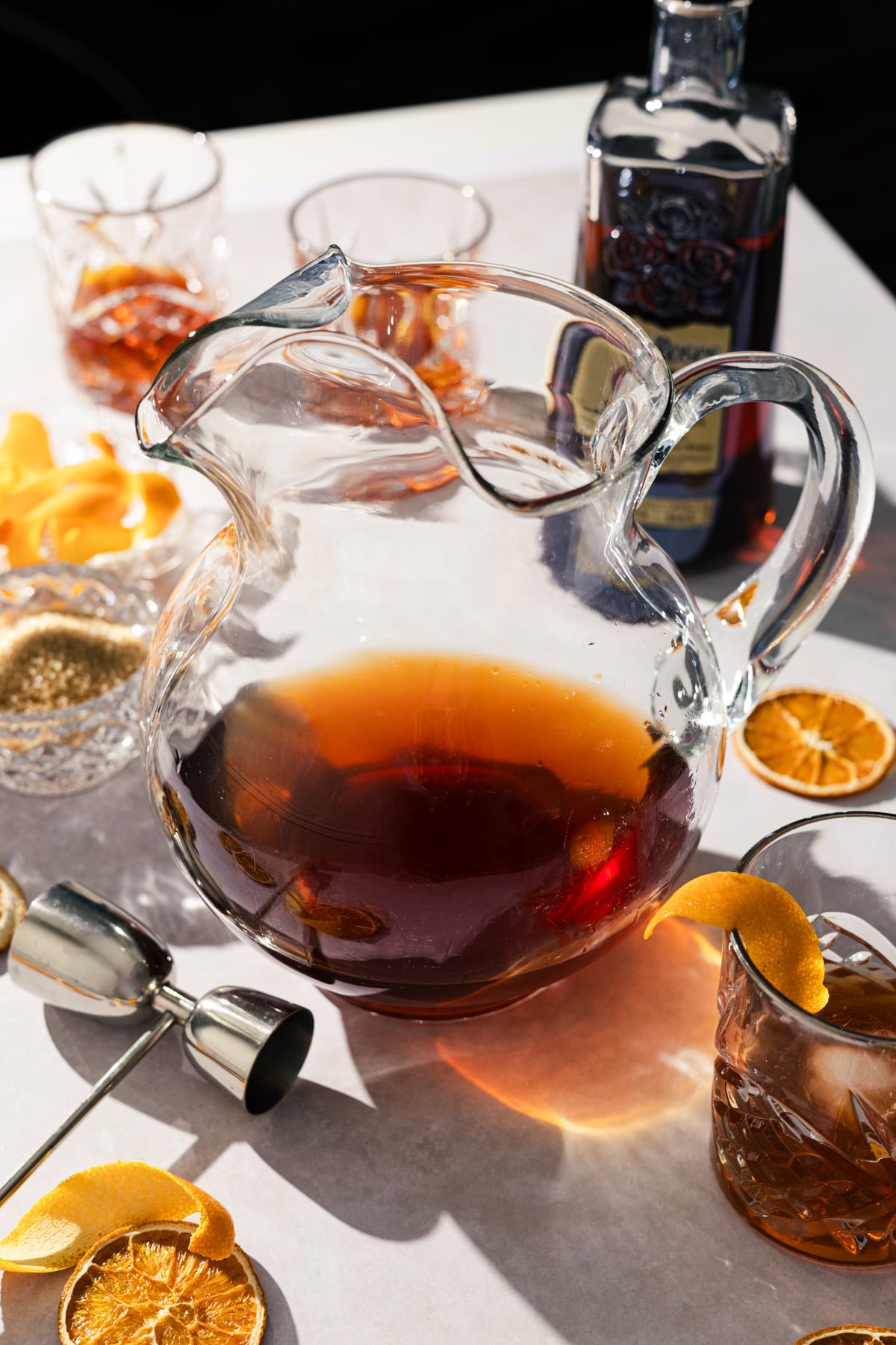 A glass pitcher filled with dark brown liquid sits on a table, set for a large batch Old Fashioned. Cocktail glasses, orange peels, dried orange slices, liquor, a jigger, and sugar await as sunlight casts soft shadows.