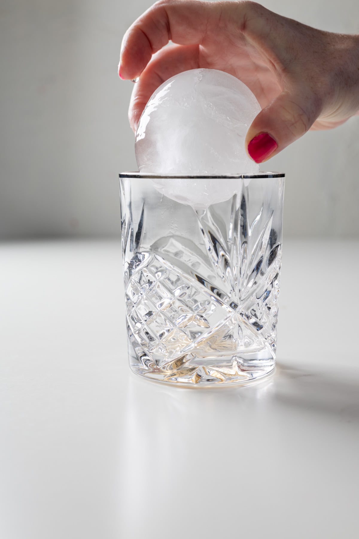 A hand with red nail polish places a large round ice sphere into a faceted, empty glass-perfect for serving a large batch old fashioned-on a white surface.
