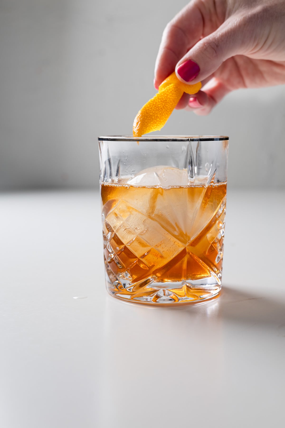 A hand garnishes a glass of amber-colored large batch Old Fashioned with an orange peel. The drink contains large ice cubes and is served in a faceted glass on a white surface.
