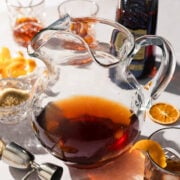 A glass pitcher filled with a dark brown liquid sits on a table surrounded by cocktail-making items, perfect for serving a large batch old fashioned, including a jigger, glasses, orange peels, dried orange slice, sugar bowl, and bottle of liquor.