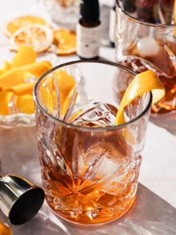 A glass of amber large batch old fashioned with ice and an orange twist sits on a marble surface. Nearby are a jigger, a bowl of orange peels, dried orange slices, and a small dropper bottle. Sunlight casts inviting shadows.