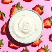 A bowl of creamy white yogurt is placed on a pink surface, surrounded by halved strawberries with green stems.