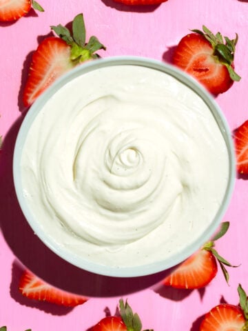 A bowl of creamy white yogurt is placed on a pink surface, surrounded by halved strawberries with green stems.