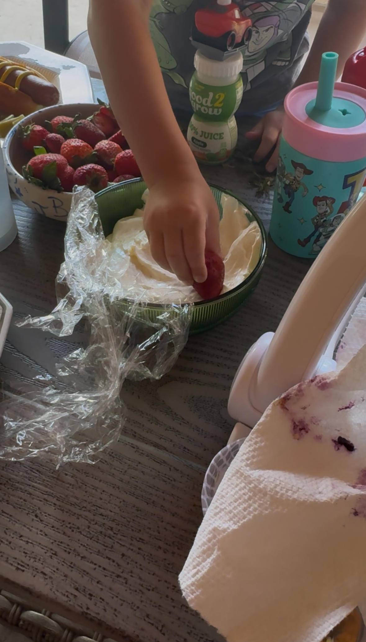 A child's hand dips a strawberry into a bowl of 5 minute marshmallow fluff fruit dip, partially covered with plastic wrap. Fresh strawberries, juice, and a blue kids cup with cartoon characters are on the table.