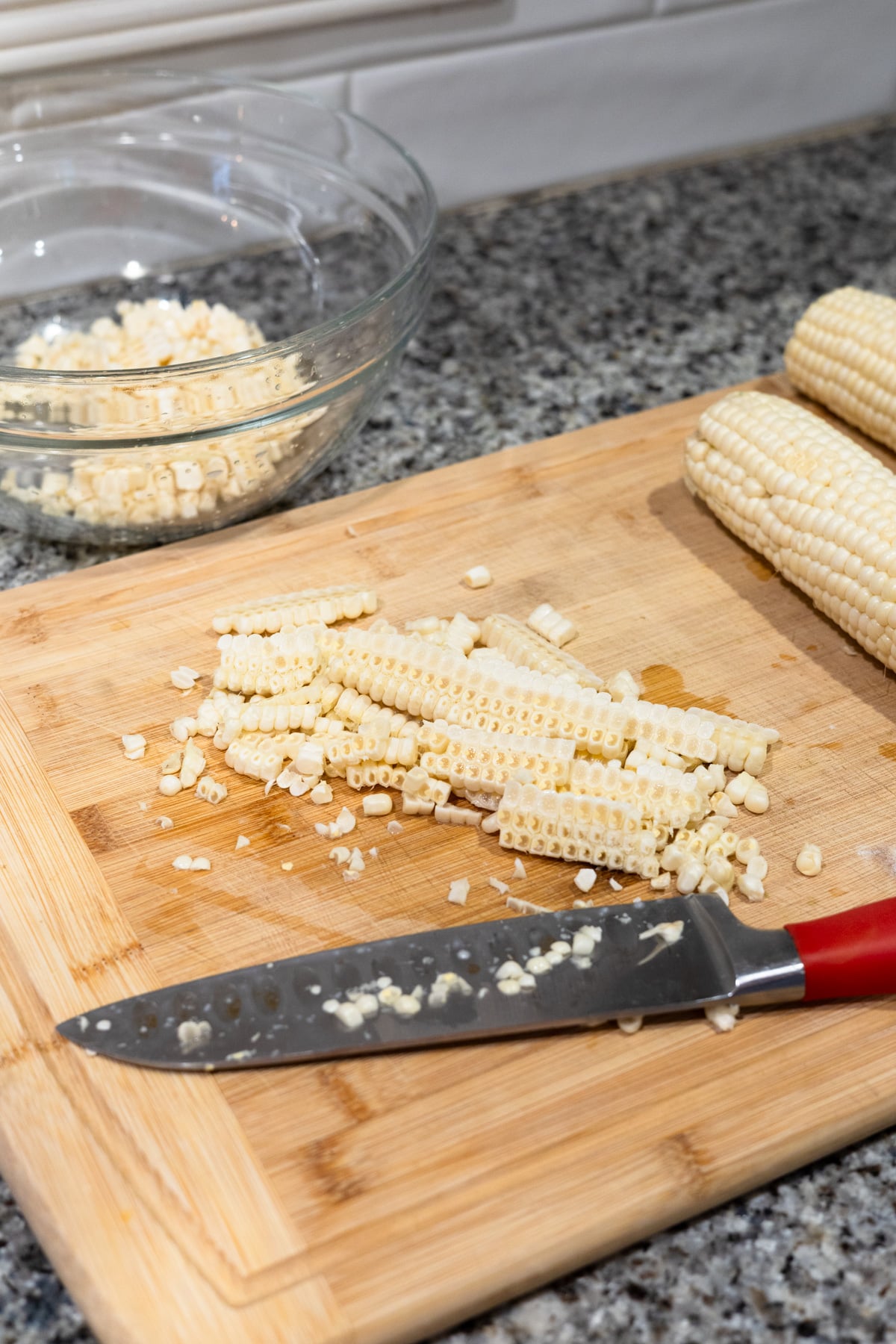 Corn kernels and cobs on a wooden cutting board next to a knife with a red handle; a glass bowl with more kernels is in the background on a granite countertop.