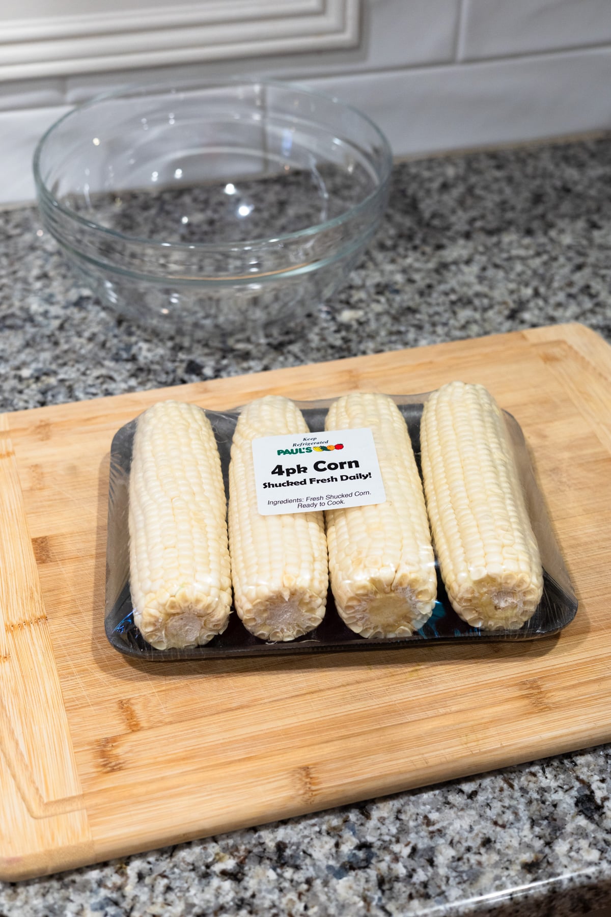 A package of four husked corn cobs sits on a wooden cutting board, with a glass mixing bowl in the background on a granite kitchen countertop.