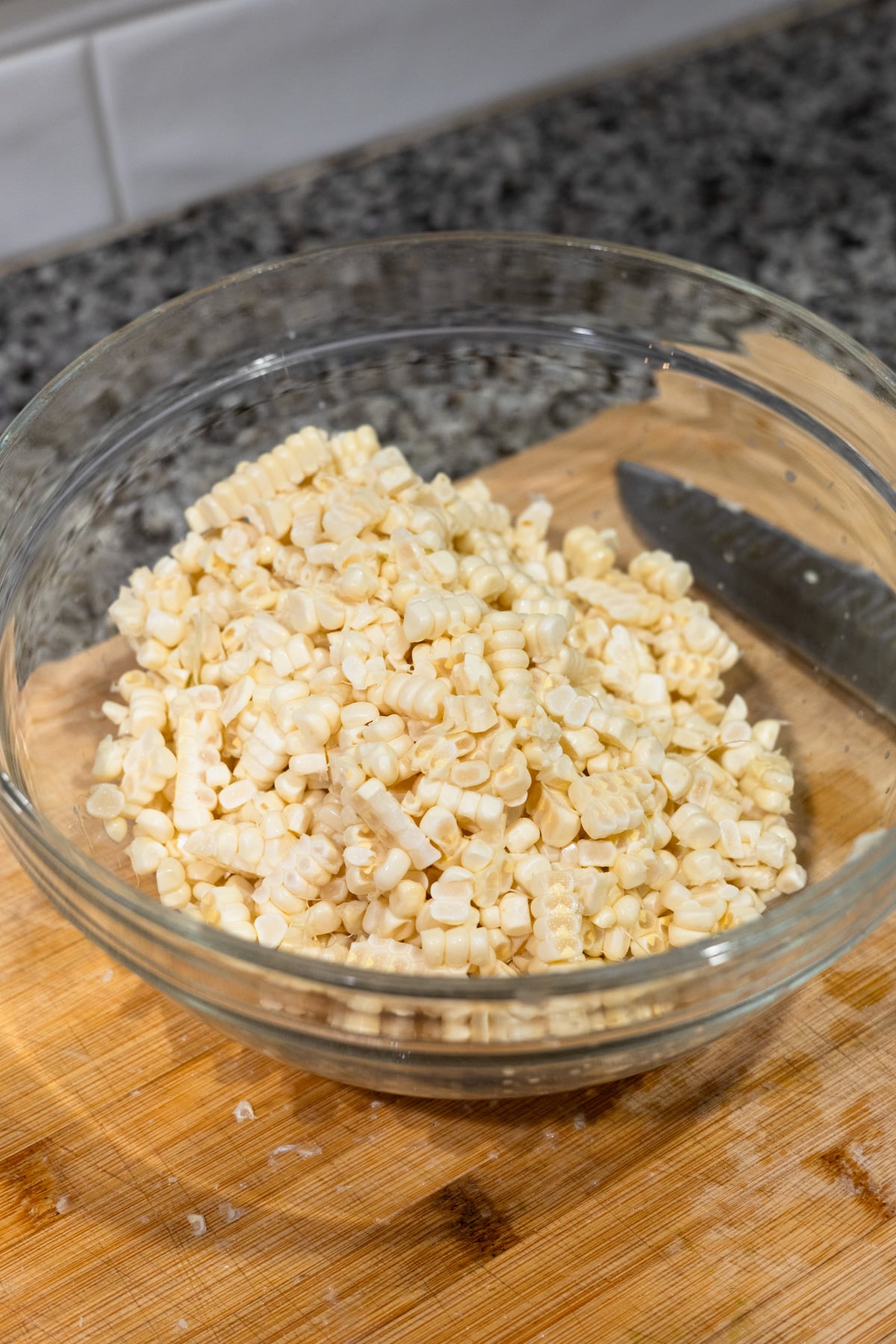 A glass bowl filled with freshly cut corn kernels sits on a wooden cutting board next to a knife, with a granite countertop and white tile backsplash in the background.