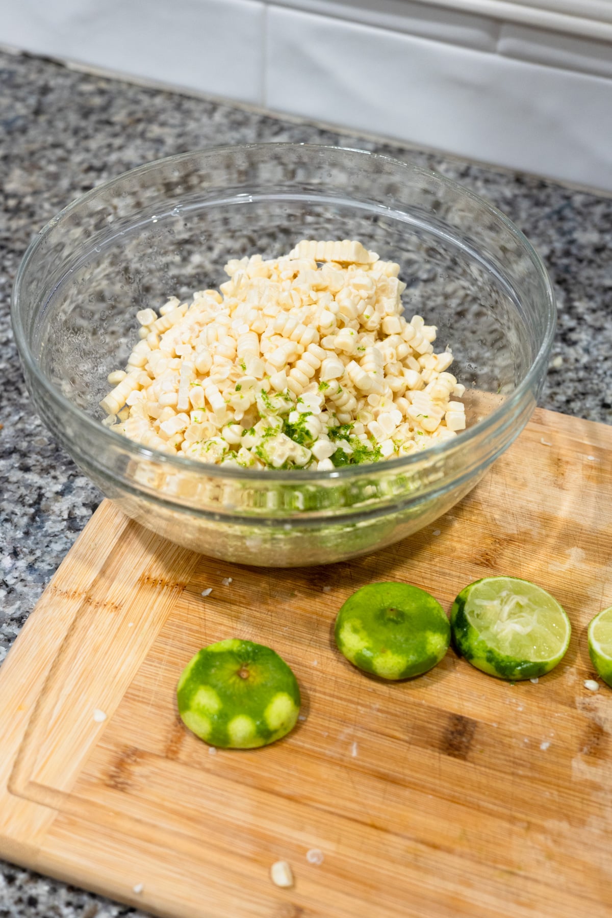 A glass bowl filled with corn kernels and lime zest sits on a granite countertop. Next to it, a wooden cutting board holds sliced and squeezed limes.
