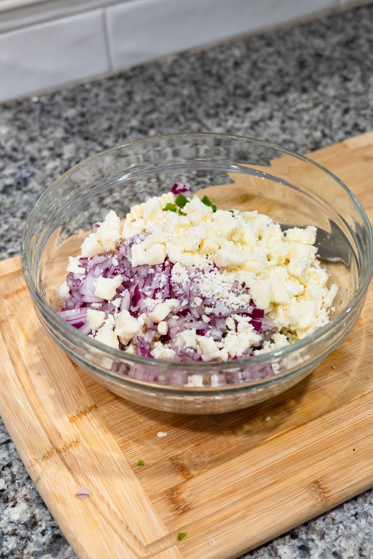 A glass bowl on a wooden cutting board contains chopped red onions, crumbled white cheese, and herbs. The bowl is on a granite countertop with a tiled backsplash in the background.