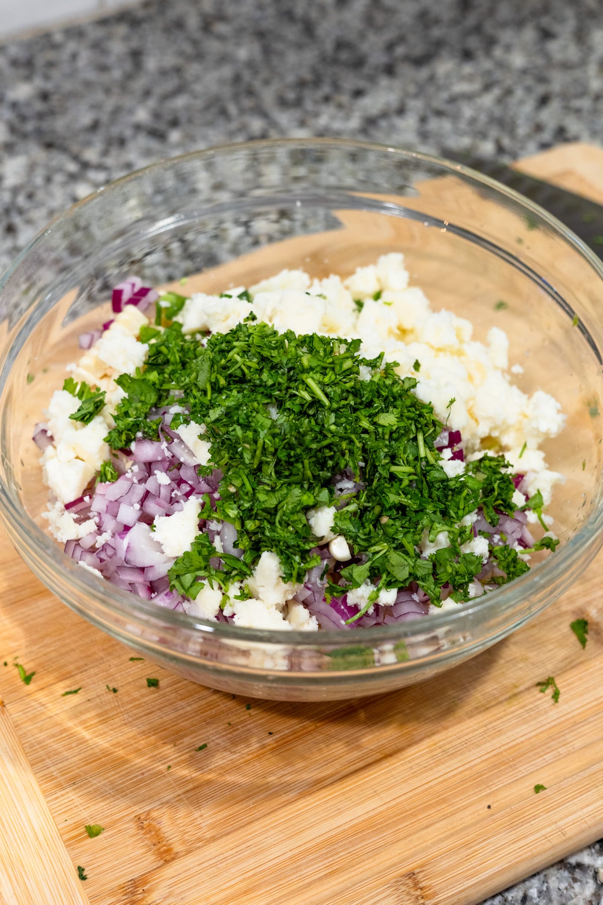 A glass bowl on a wooden cutting board contains chopped red onion, crumbled white cheese, and freshly chopped cilantro, ready to be mixed. The background shows a granite countertop.
