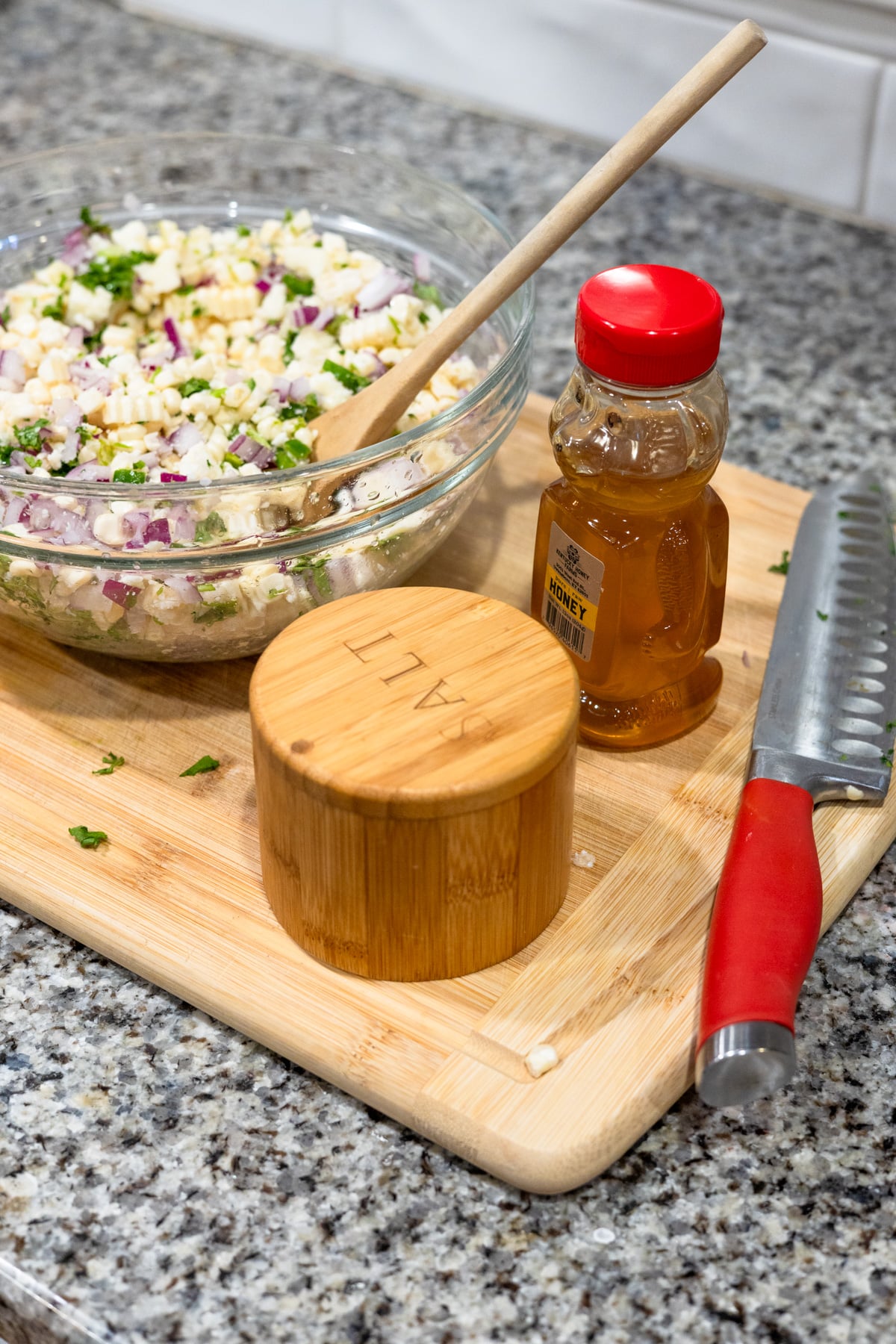 A glass bowl of corn salad with a wooden spoon sits on a cutting board, next to a container labeled SALT, a small honey bottle, and a knife with a red handle on a granite countertop.