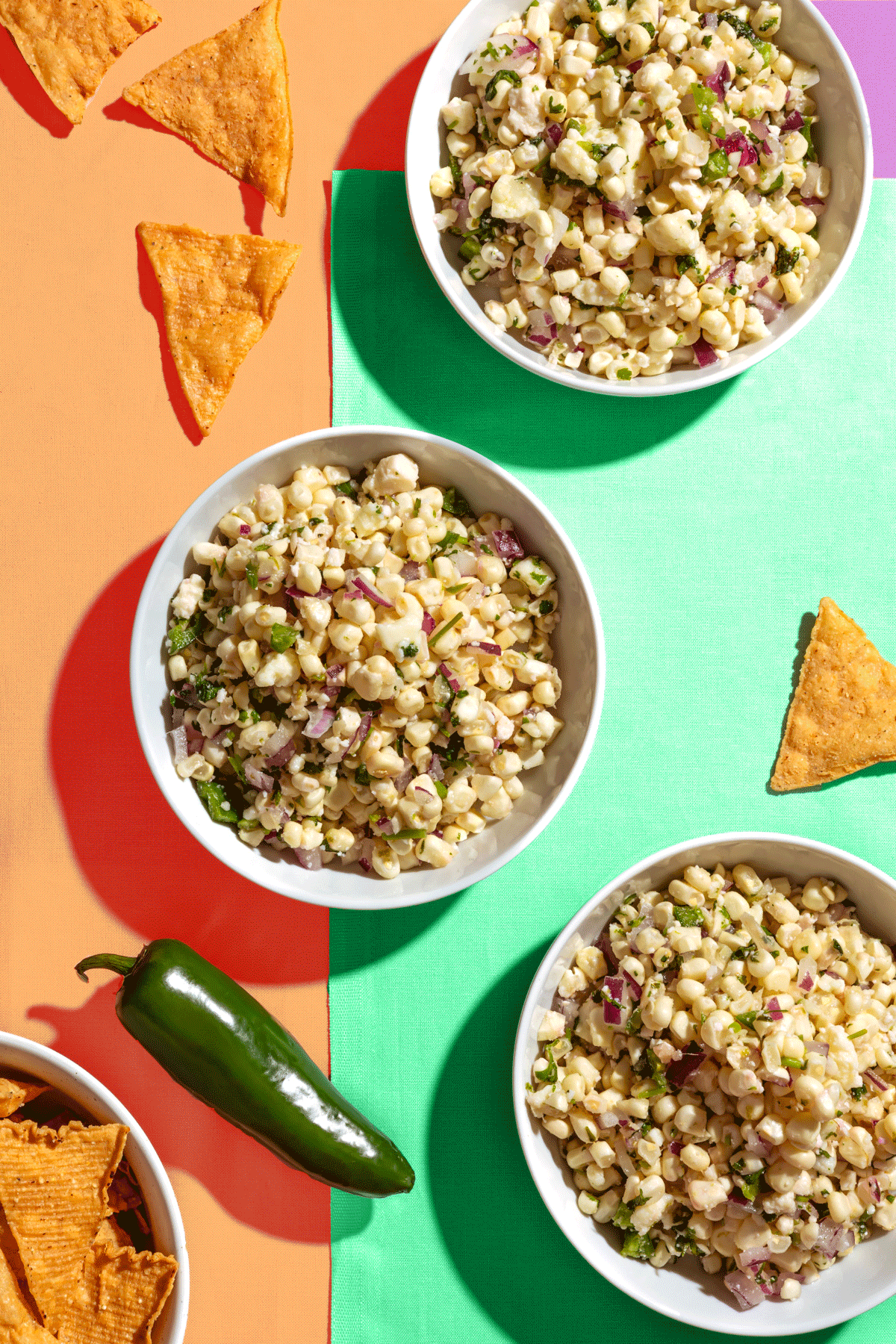 Three bowls of corn salad with diced red onion and herbs are arranged on a colorful, geometric background. Tortilla chips and a green jalapeño pepper are also on the table, with chips scattered around.