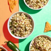 Three bowls of corn salad with chopped red onion and herbs on a colorful orange and green surface, surrounded by tortilla chips and a jalapeño pepper.