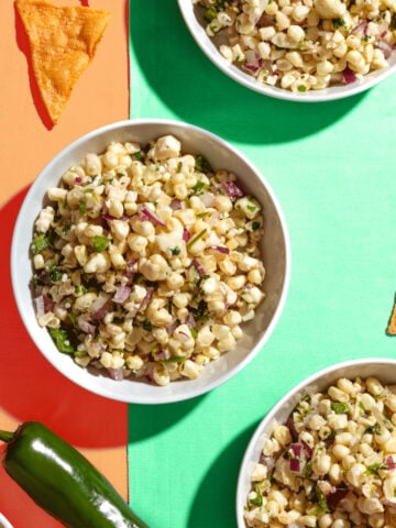Three bowls of corn salad with chopped red onion and herbs on a colorful orange and green surface, surrounded by tortilla chips and a jalapeño pepper.