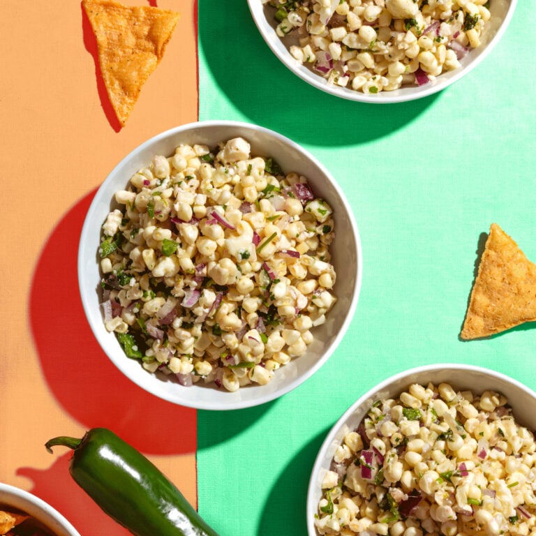 Three bowls of corn salad with chopped red onion and herbs on a colorful orange and green surface, surrounded by tortilla chips and a jalapeño pepper.