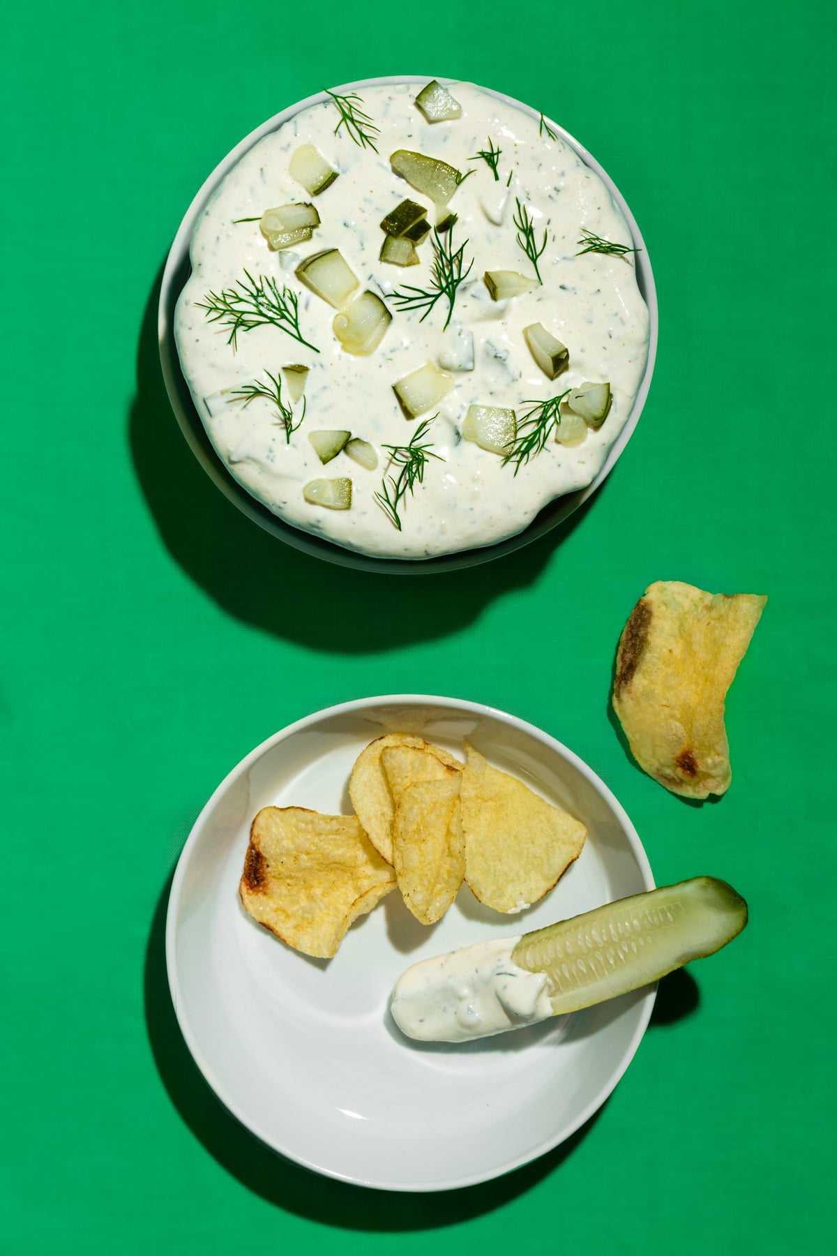 A bowl of creamy dip topped with chopped pickles and dill sits above a plate with potato chips, a pickle spear, and dip on a bright green background.
