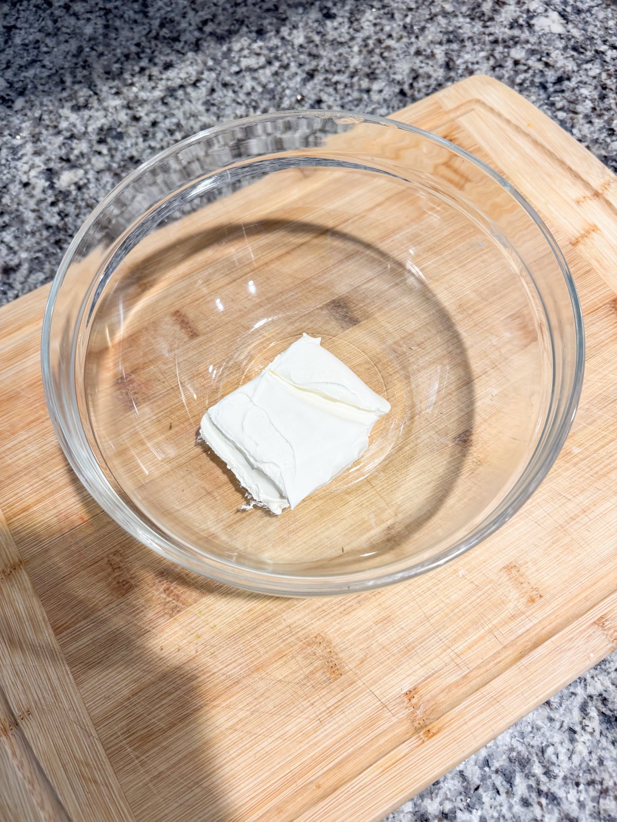 A clear glass bowl containing a small block of cream cheese sits on a wooden cutting board atop a speckled gray countertop.