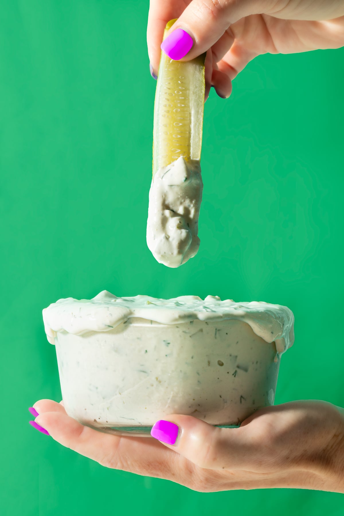 A hand with purple nail polish dips a pickle spear into a bowl of creamy ranch dip against a green background.