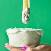 A hand with bright purple nail polish holds a glass bowl overflowing with creamy dip, as a celery stick coated in dip is lifted above the bowl against a vibrant green background.